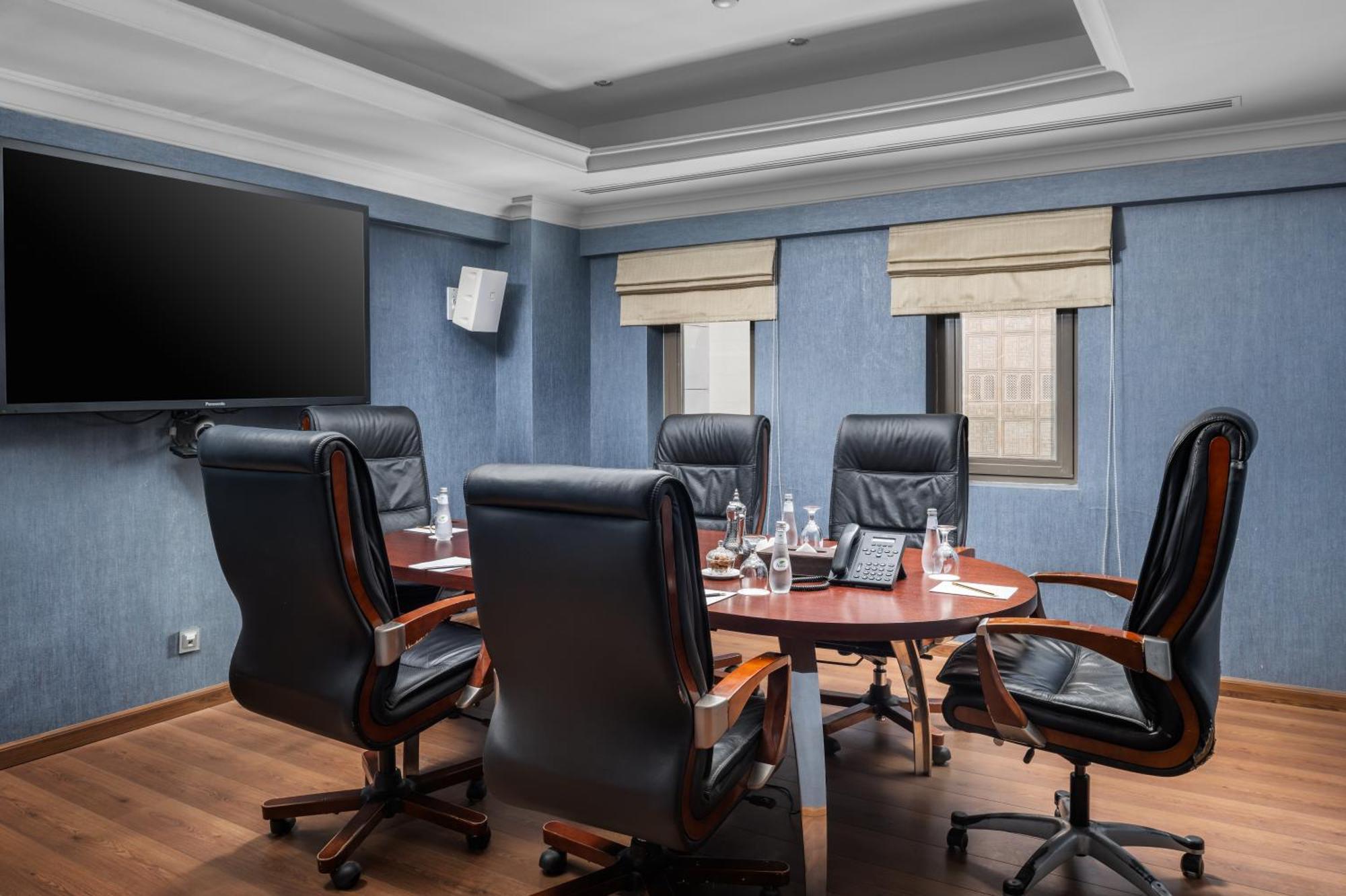 Al Aqeeq Madinah Hotel Médine Extérieur photo The photo shows a modern conference room. It features a round wooden table surrounded by several black leather swivel chairs. The walls have a blue color scheme, and there is a large flat-screen TV mounted on one wall. The room has two windows with b