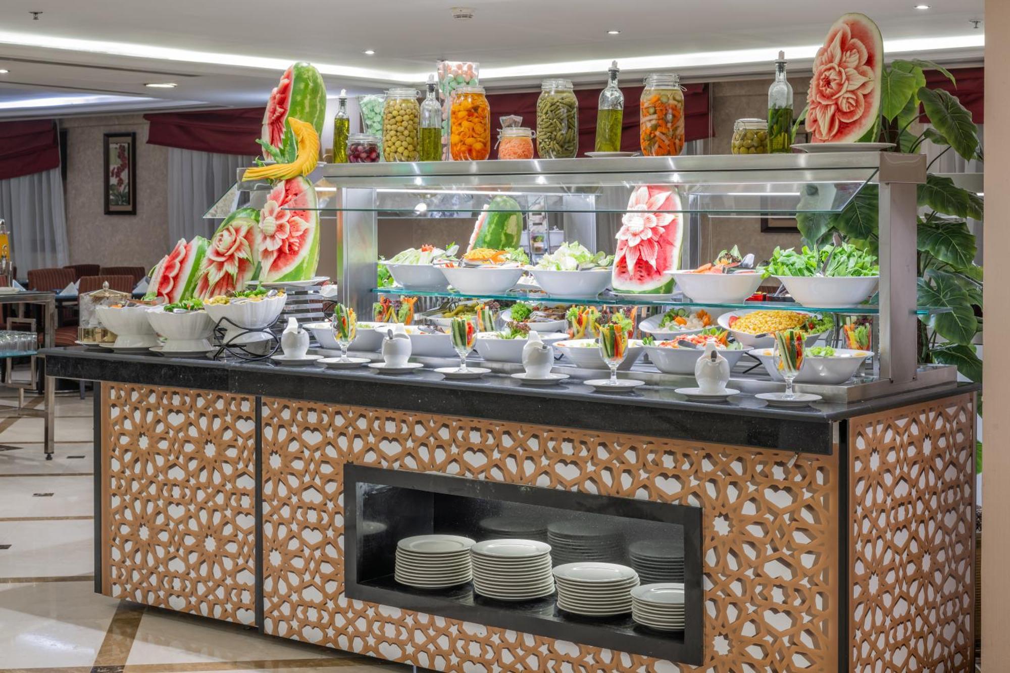 Al Aqeeq Madinah Hotel Médine Extérieur photo The photo shows a buffet display that features an array of colorful food items. In the foreground, there are several large bowls filled with various salads and dishes. The presentation is visually appealing, with decorative watermelon carvings and an