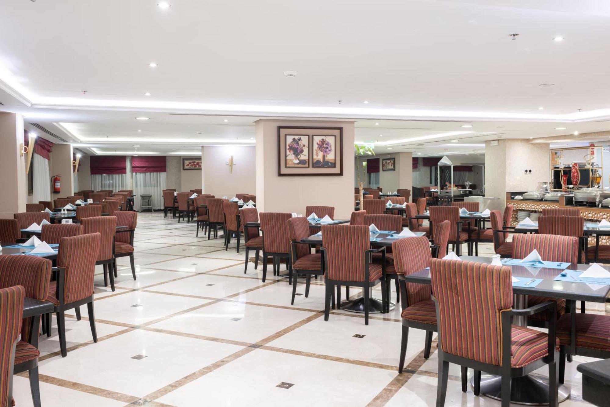 Al Aqeeq Madinah Hotel Médine Extérieur photo The photo shows the interior of a spacious restaurant. It features a well-organized dining area with neatly arranged tables and chairs. The tables are dressed with blue napkins, and there is a light-colored flooring with a grid pattern. The walls are