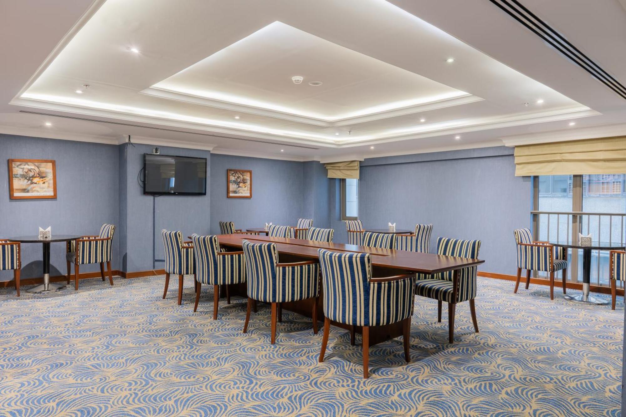 Al Aqeeq Madinah Hotel Médine Extérieur photo The photo shows a spacious conference or meeting room. It features a light blue color scheme with a patterned carpet. There are several rectangular tables arranged in a group, surrounded by striped chairs. The room is well-lit with modern ceiling lig