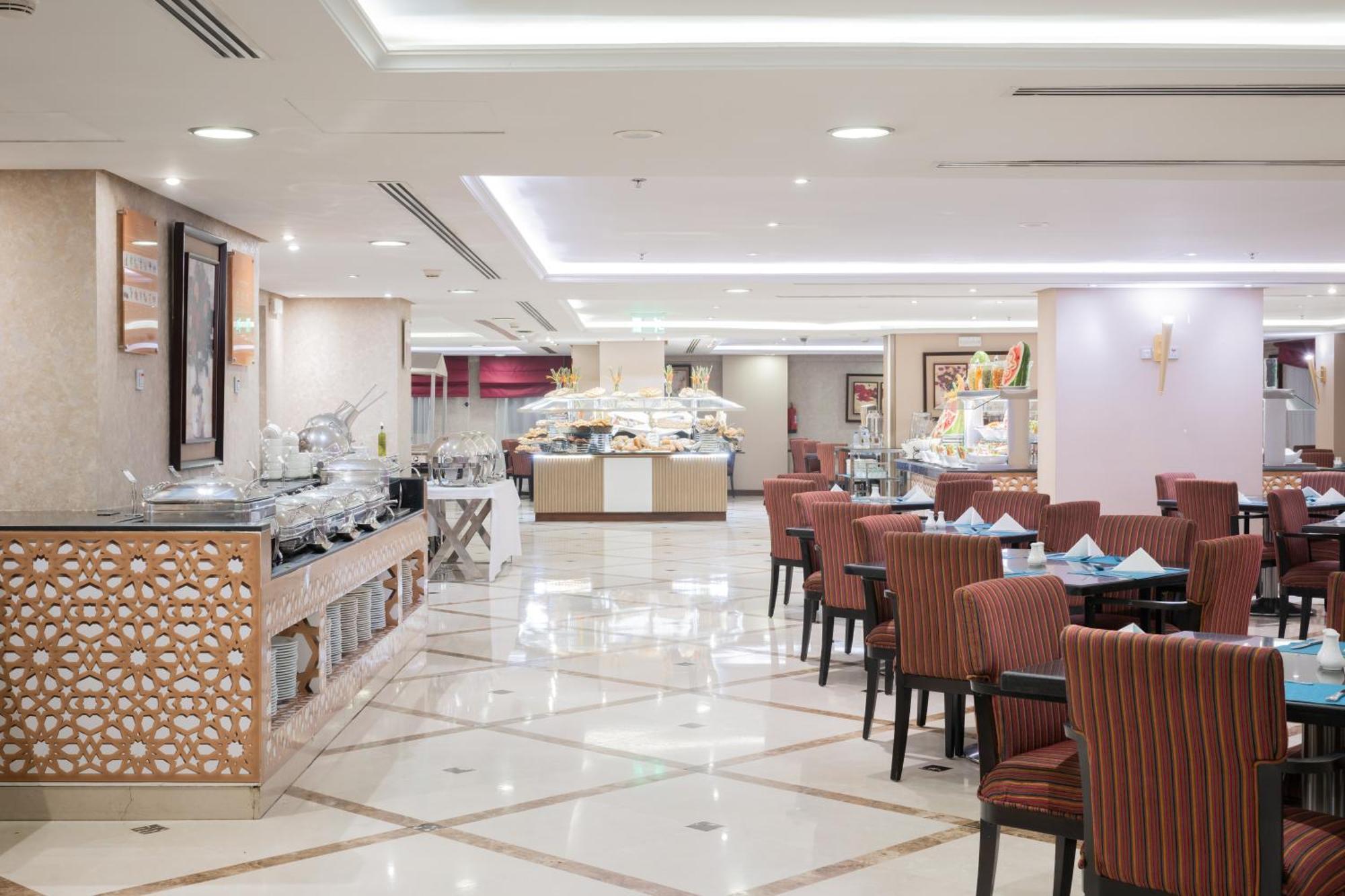 Al Aqeeq Madinah Hotel Médine Extérieur photo The photo shows an elegantly designed restaurant or dining area. It features a spacious layout with a glossy tiled floor and bright, soft lighting. There are several dining tables with striped chairs arranged neatly. Along one side, there's a buffet 