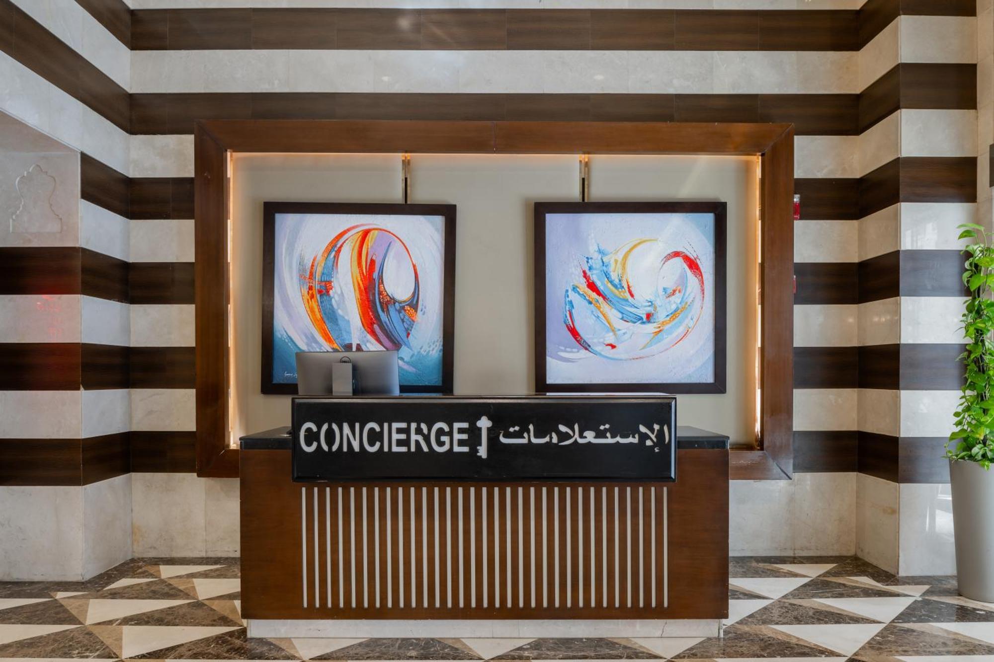 Al Aqeeq Madinah Hotel Médine Extérieur photo The image shows a concierge desk in a hotel lobby. The desk is made of wood with a striped design and features two large, colorful abstract paintings on the wall behind it. The sign on the desk reads "CONCIERGE" in English and has Arabic text below i