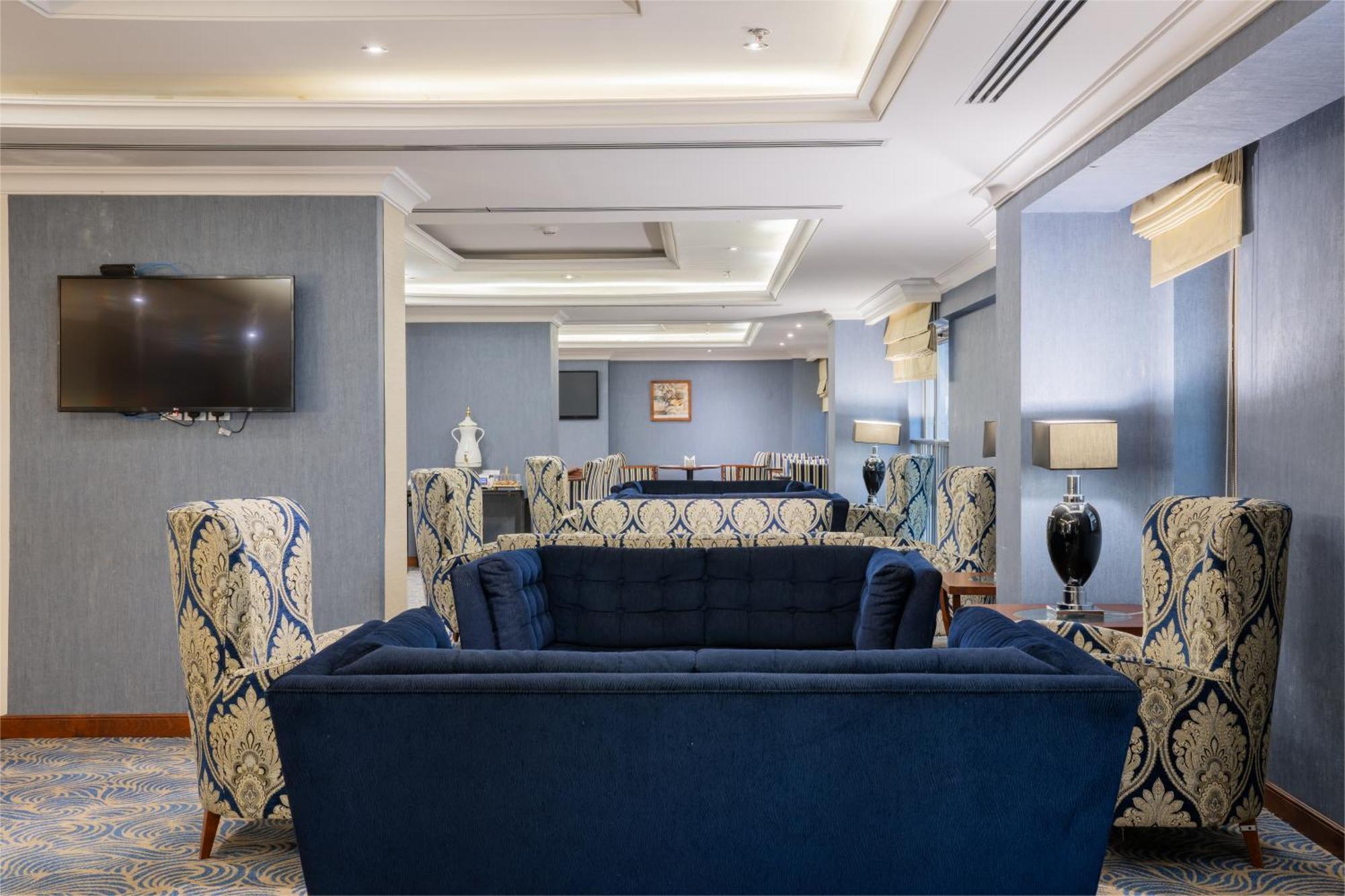 Al Aqeeq Madinah Hotel Médine Extérieur photo The photo shows a stylish and modern lounge area. There are two patterned armchairs with blue and cream designs, positioned next to a deep blue couch. The room has a light blue wall with a flat-screen TV mounted on one side. Soft lighting from stylis