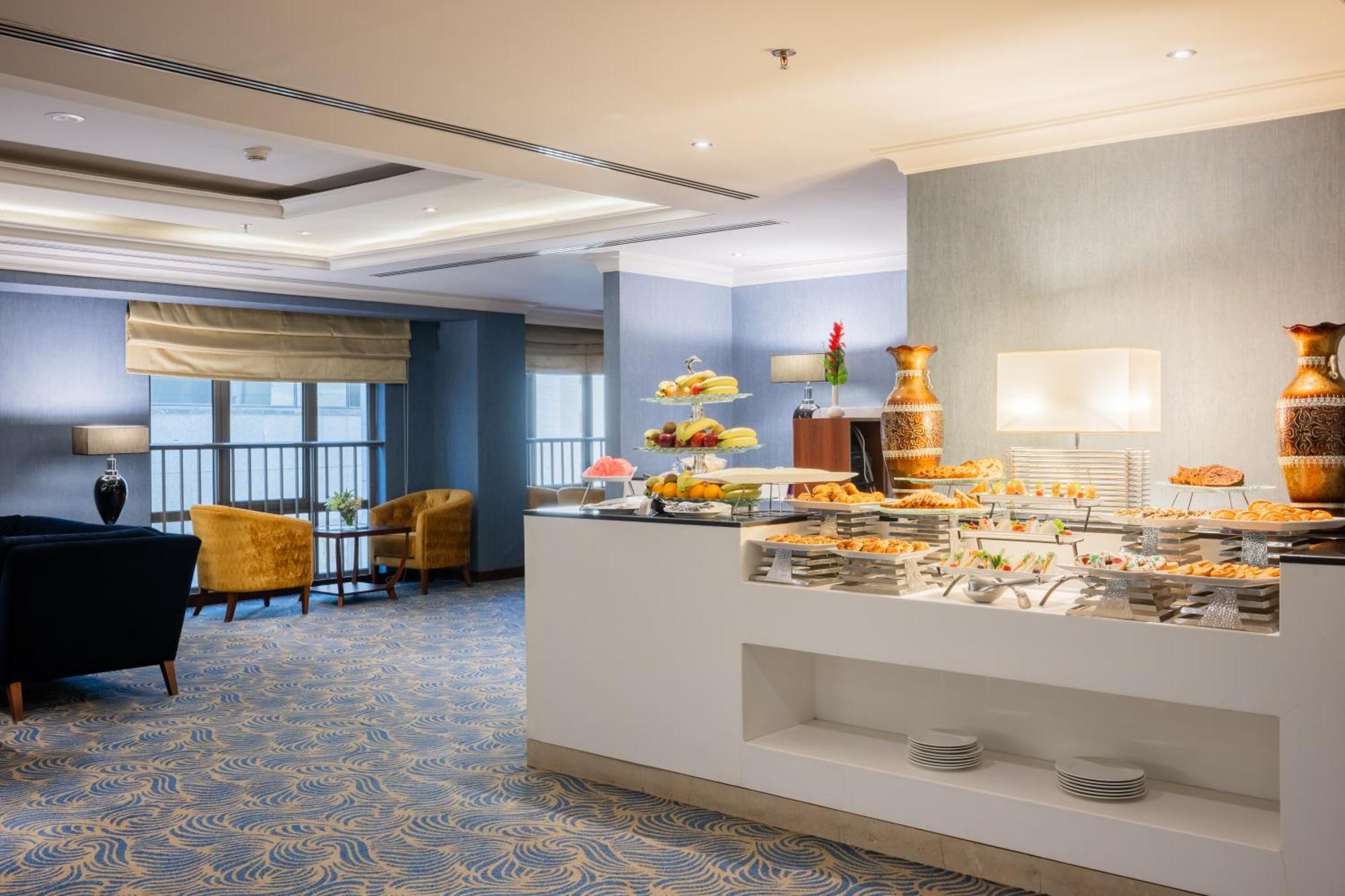 Al Aqeeq Madinah Hotel Médine Extérieur photo The photo shows a modern dining area, likely in a hotel or restaurant. On the right side, there is a buffet table with various food items displayed, including pastries and desserts. Behind the table, a decorative fountain is visible. The room feature