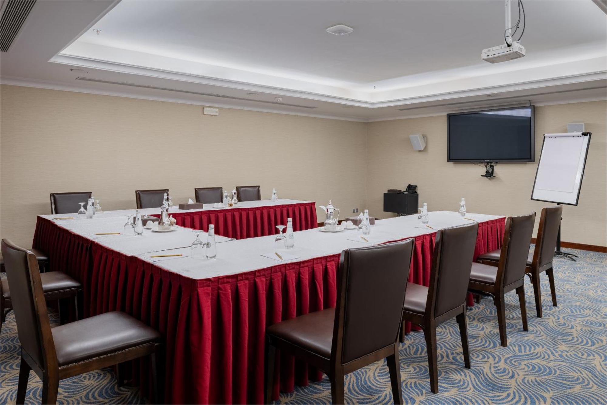 Al Aqeeq Madinah Hotel Médine Extérieur photo The image shows a conference room set up for a meeting. There is a U-shaped arrangement of tables covered with white linens and red table skirts. Each table position is equipped with a water bottle and a small notepad, suggesting that it is designed 