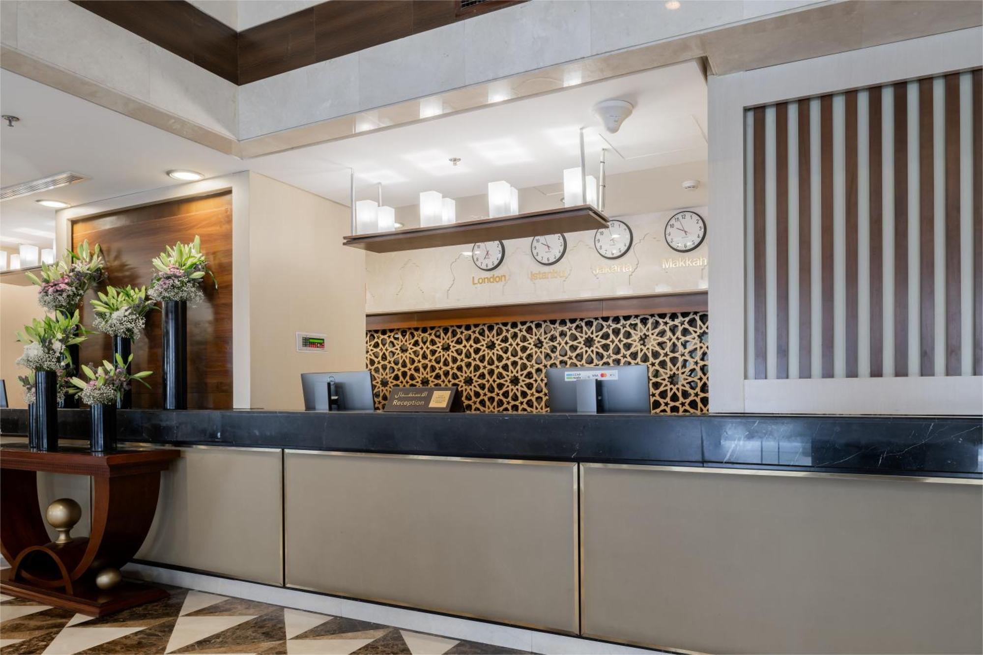 Al Aqeeq Madinah Hotel Médine Extérieur photo The photo shows the interior of a hotel reception area. The reception desk is designed with a sleek, modern look featuring a dark countertop and a light-colored front. Above the desk, there are several clocks displaying different times, suggesting an