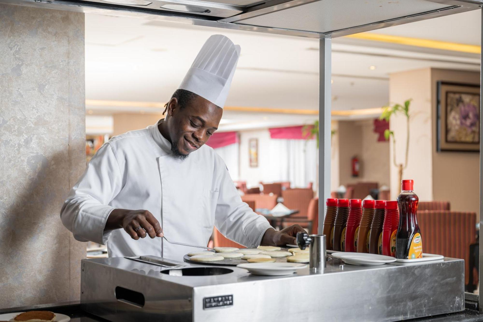 Al Aqeeq Madinah Hotel Médine Extérieur photo The photo shows a chef in a professional kitchen setting. He is wearing a traditional white chef's uniform and a tall white chef's hat, and he appears to be smiling while cooking. The chef is standing at a stainless steel cooking station, where he is