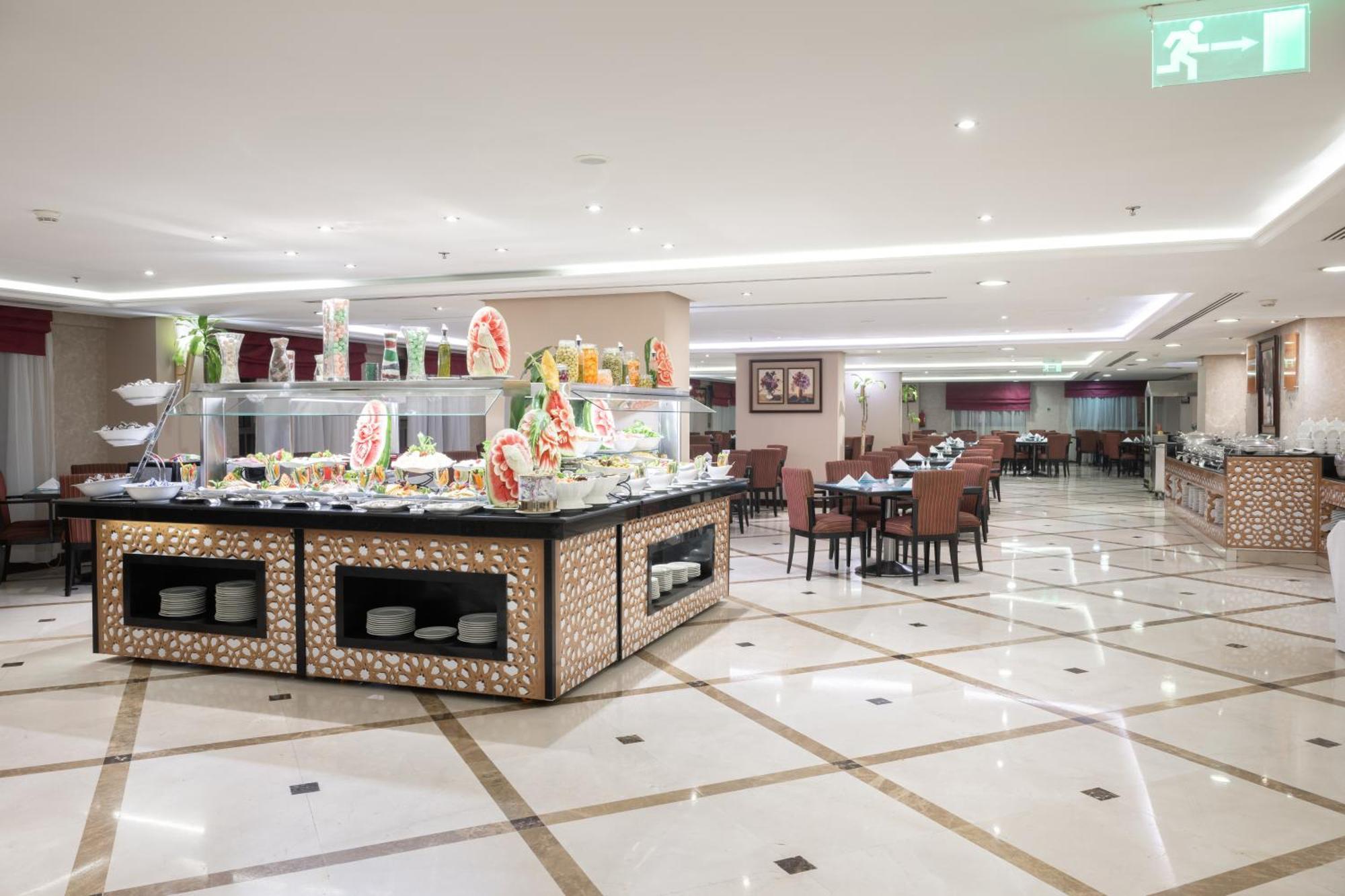 Al Aqeeq Madinah Hotel Médine Extérieur photo The photo shows a spacious restaurant interior with a modern design. In the foreground, there is a large buffet counter filled with various food items, beautifully arranged and decorated. The counter features a backlit display, showcasing fruits and 