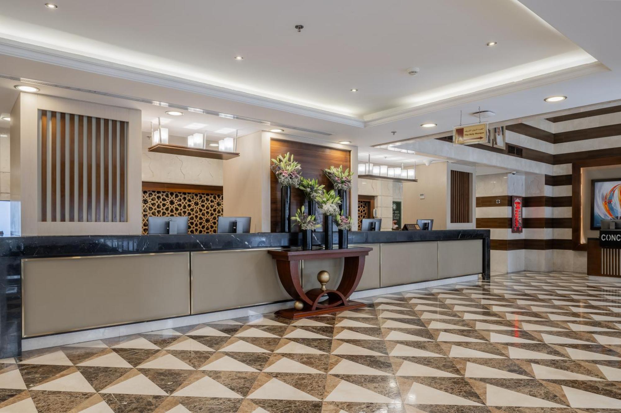 Al Aqeeq Madinah Hotel Médine Extérieur photo The photo depicts a hotel reception area. The check-in counter is long and sleek, with a smooth surface and decorative elements. Behind the counter, there are several computer monitors. The walls feature stylish designs, including wooden panels and g