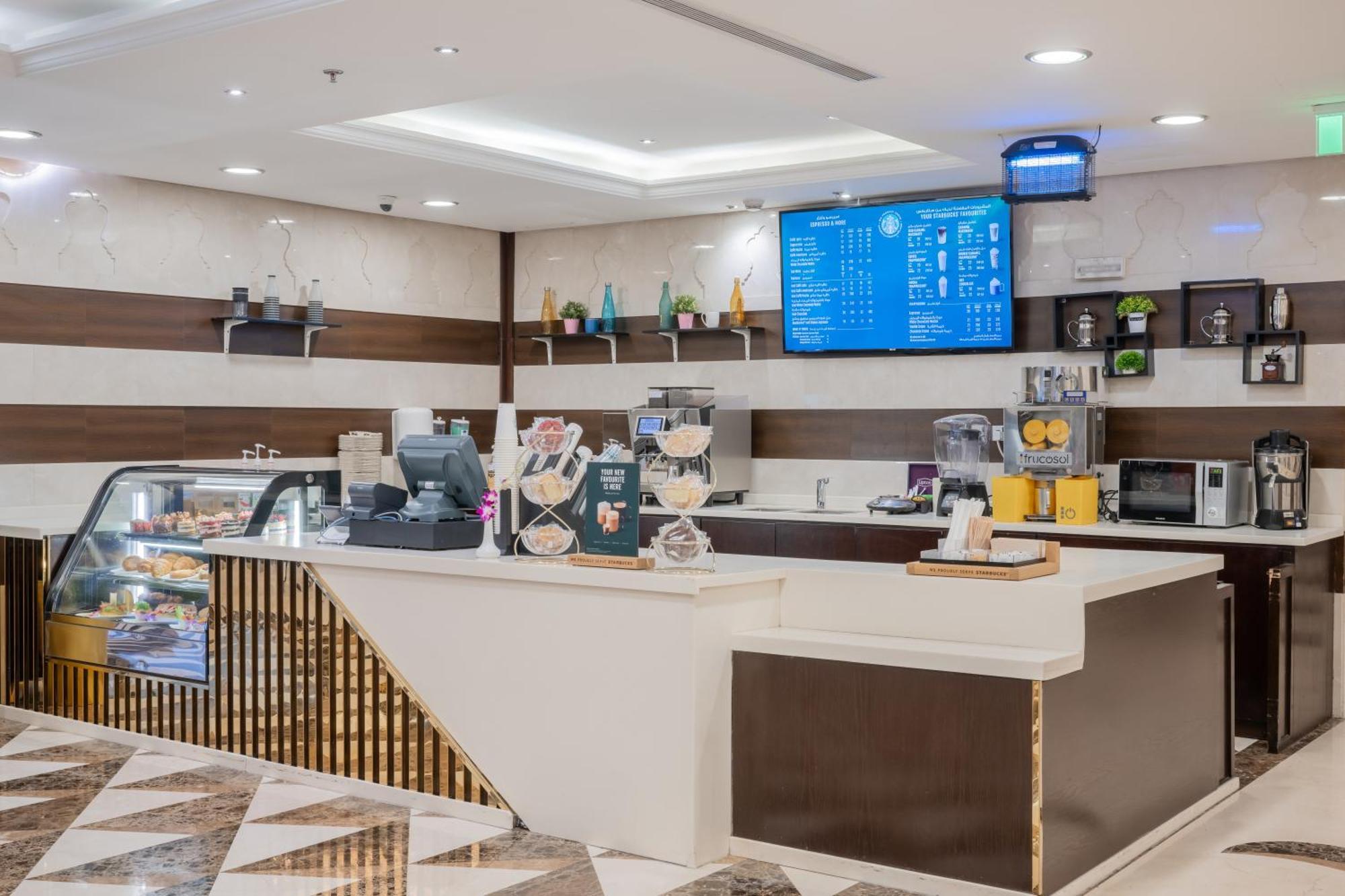 Al Aqeeq Madinah Hotel Médine Extérieur photo The photo shows a modern café or coffee shop interior. There's a counter area made of smooth white and dark brown materials, featuring a cash register and display items. Behind the counter, shelves are stocked with various beverage items and equipmen
