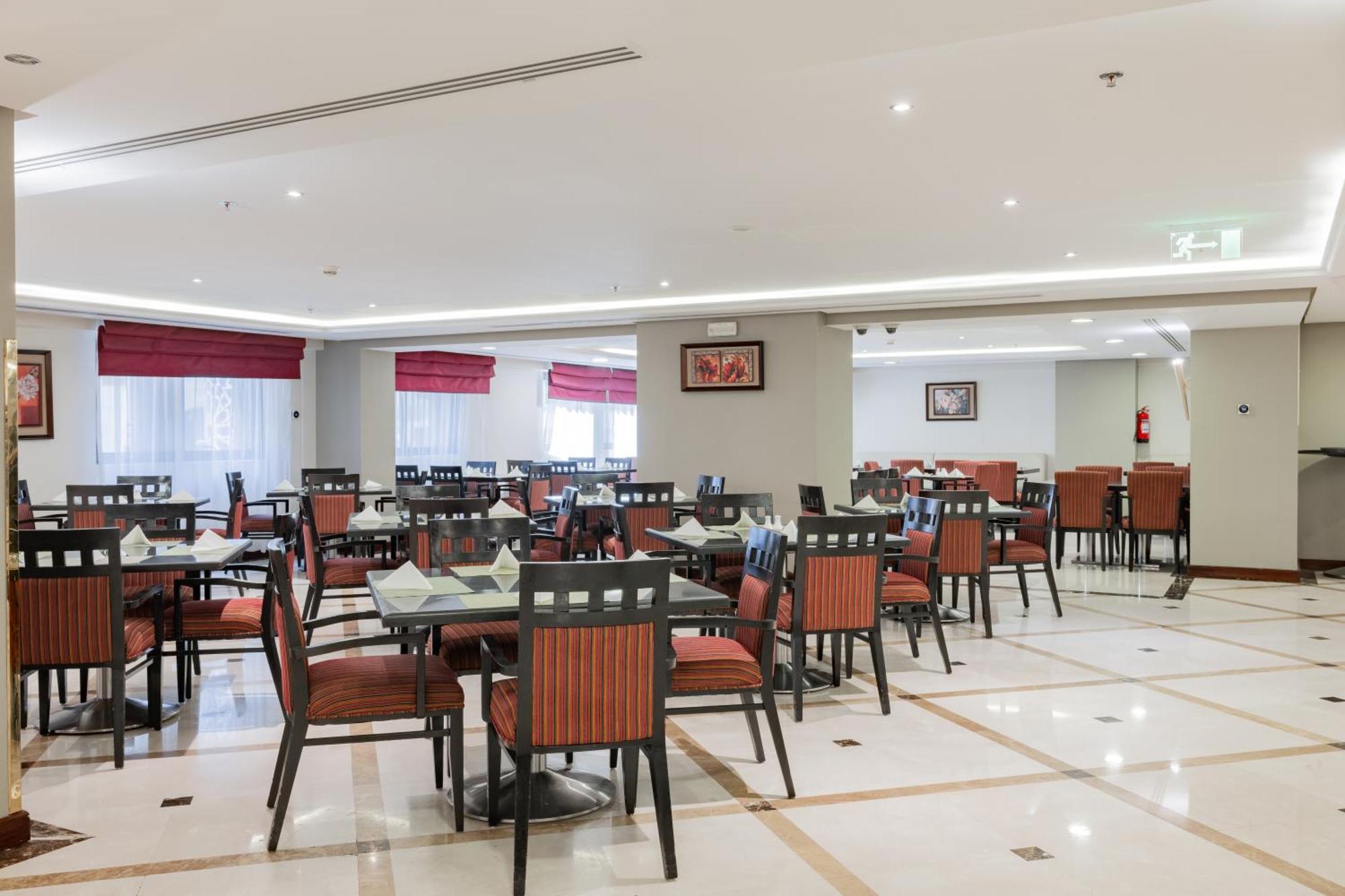 Al Aqeeq Madinah Hotel Médine Extérieur photo The photo shows the interior of a restaurant or dining area. The space features several tables arranged with chairs, most of which have striped orange and black upholstery. The flooring is likely marble or tile, and the lighting is bright, creating a
