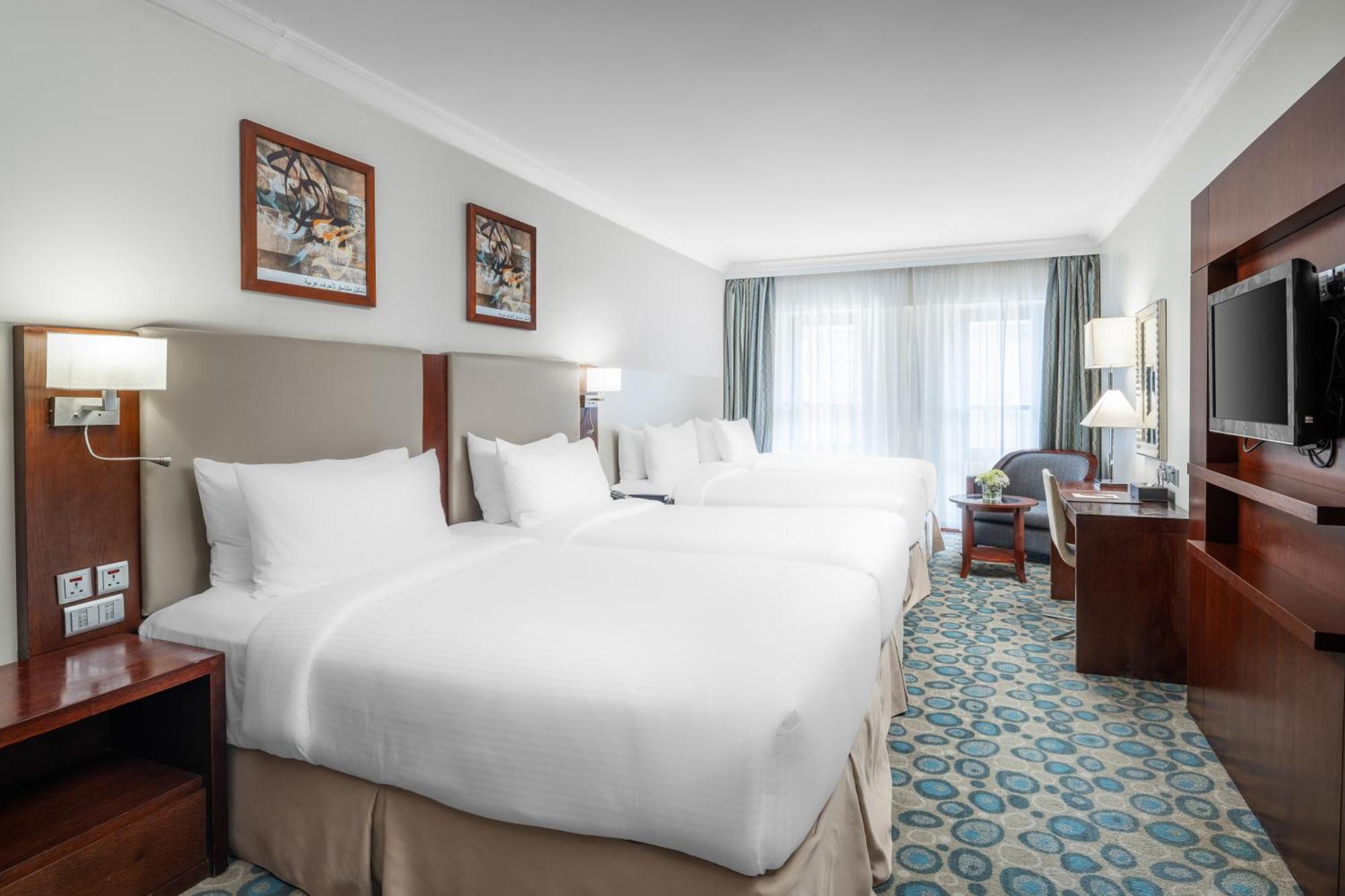 Al Aqeeq Madinah Hotel Médine Extérieur photo The photo shows a hotel room featuring a modern, clean design. There are two large beds with white linens positioned side by side. Above the beds are framed artworks, and soft lighting is provided by wall sconces. To the right, there is a wooden ente