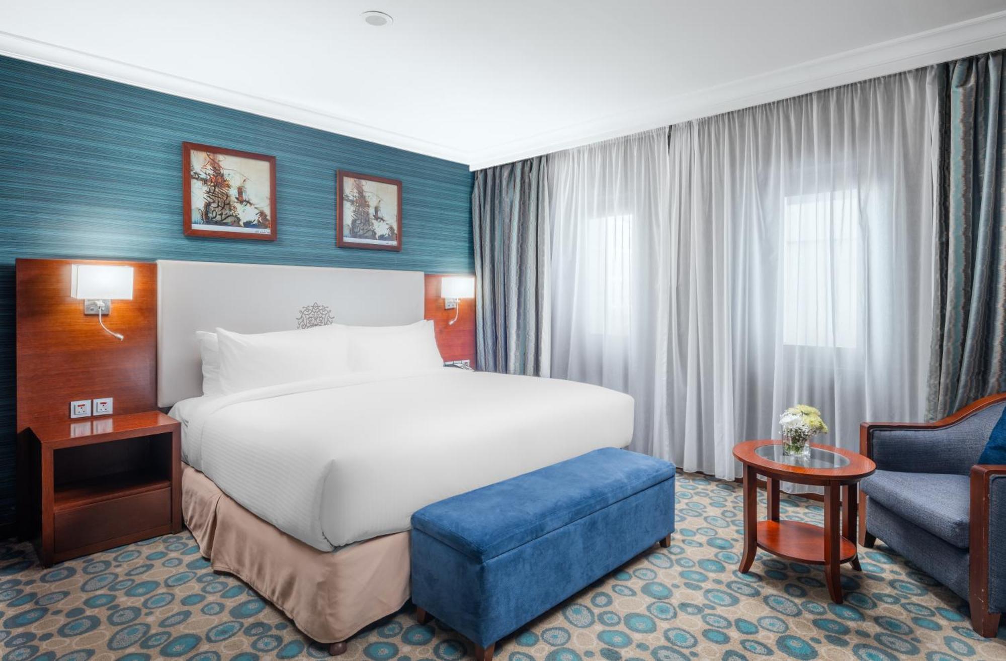 Al Aqeeq Madinah Hotel Médine Extérieur photo The photo shows a hotel room featuring a large, neatly made bed with a white duvet. The walls have a light blue accent with framed artwork above the bed. There’s a blue ottoman at the foot of the bed. To one side, there’s a small wooden table with a 