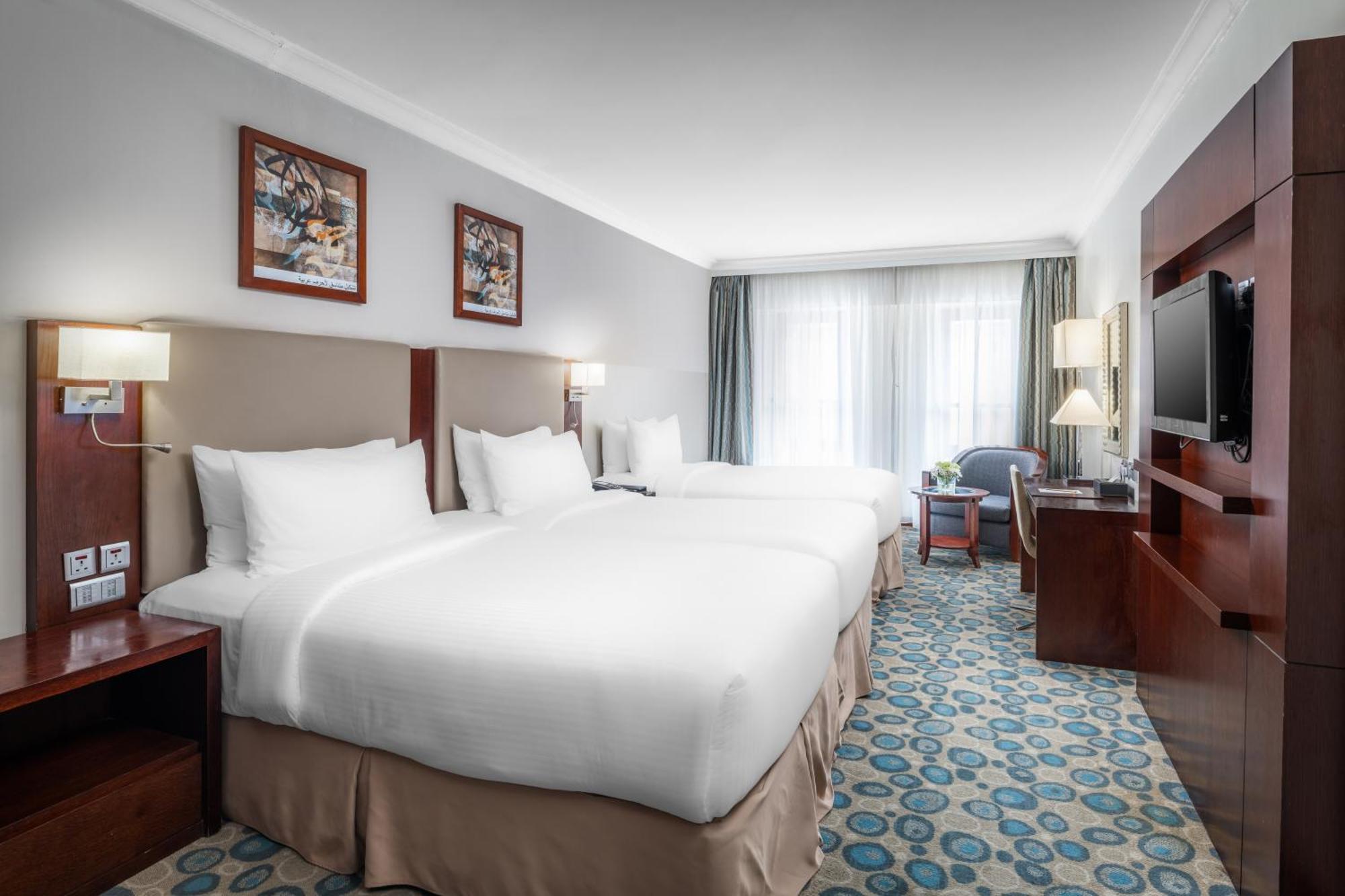 Al Aqeeq Madinah Hotel Médine Extérieur photo The photo shows a well-furnished hotel room featuring two large beds with white bedding. The walls are adorned with framed artwork. There are bedside lamps next to each bed, and a flat-screen TV is mounted on the wall. A large window allows natural l