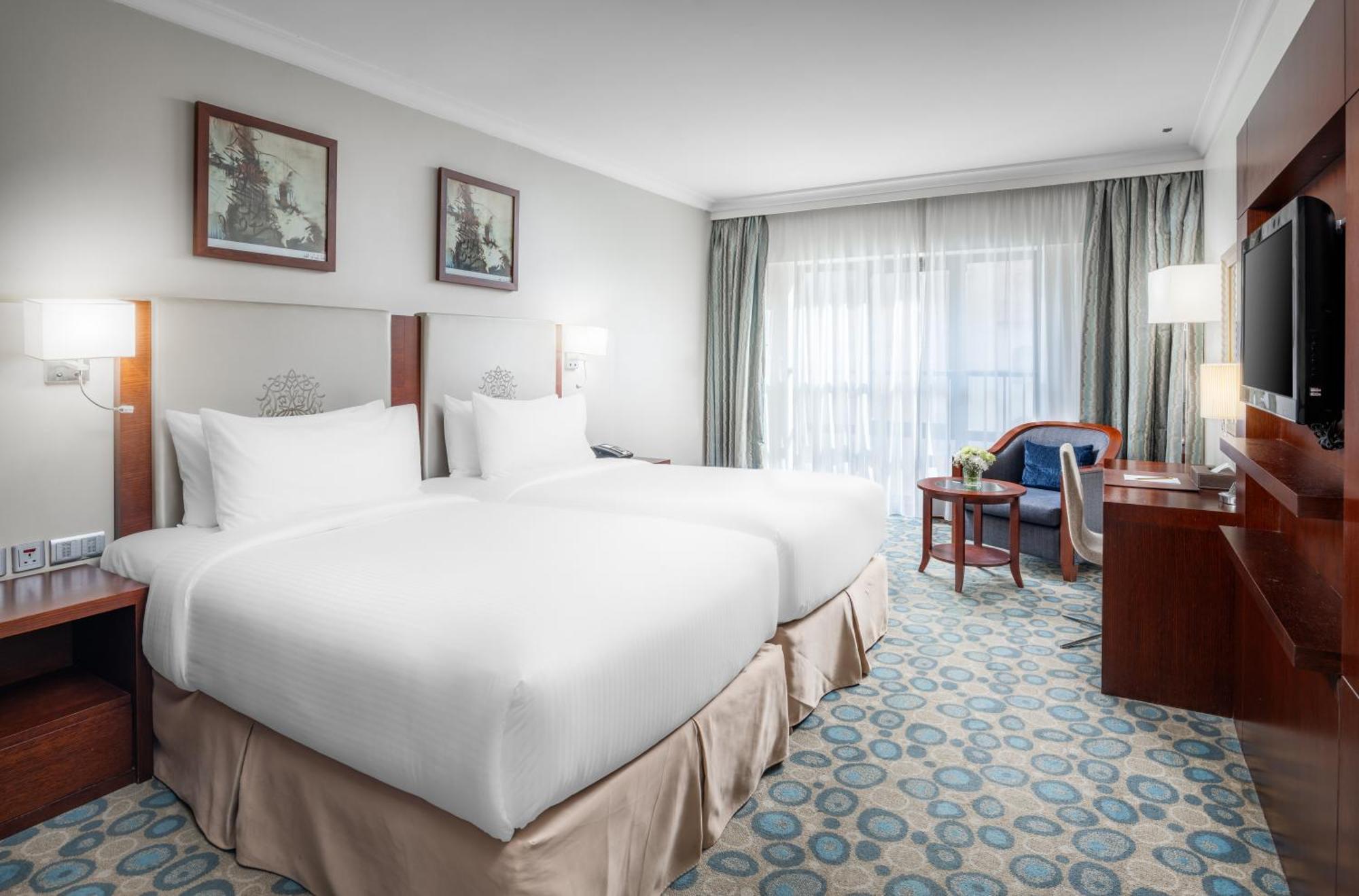 Al Aqeeq Madinah Hotel Médine Extérieur photo The photo shows a hotel room featuring two white beds with plush bedding. There are decorative artworks on the walls above the beds. A large window with sheer curtains allows natural light into the space, illuminating the room. The floor is covered w