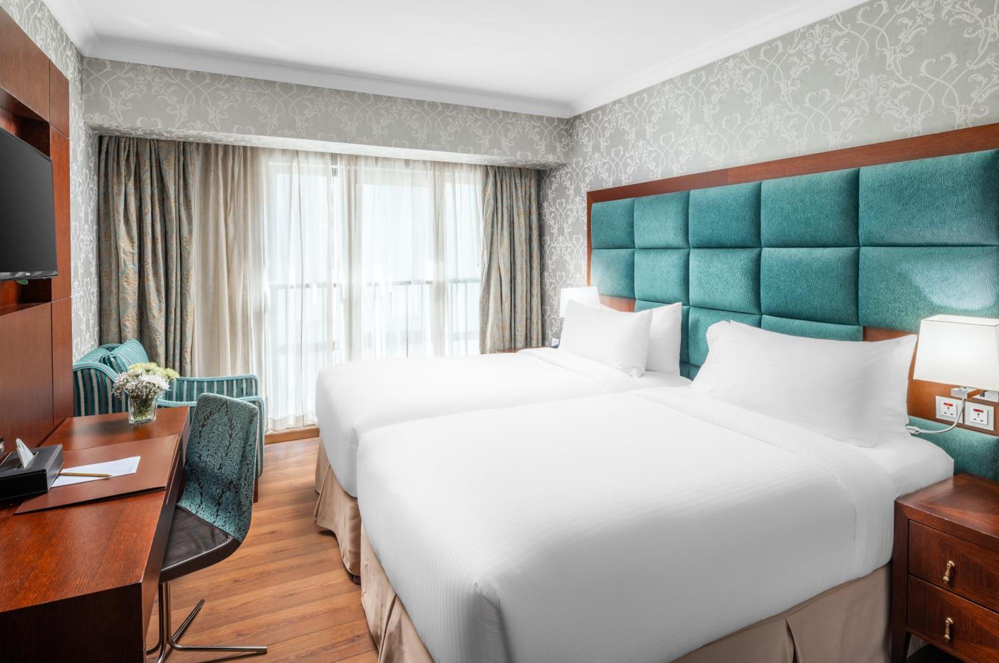 Al Aqeeq Madinah Hotel Médine Extérieur photo The photo shows a modern hotel room featuring two single beds with white bedding. The headboard is upholstered in a teal fabric. There is a wooden desk and a chair near a window that is covered with sheer curtains and drapes. A cozy armchair in a mat