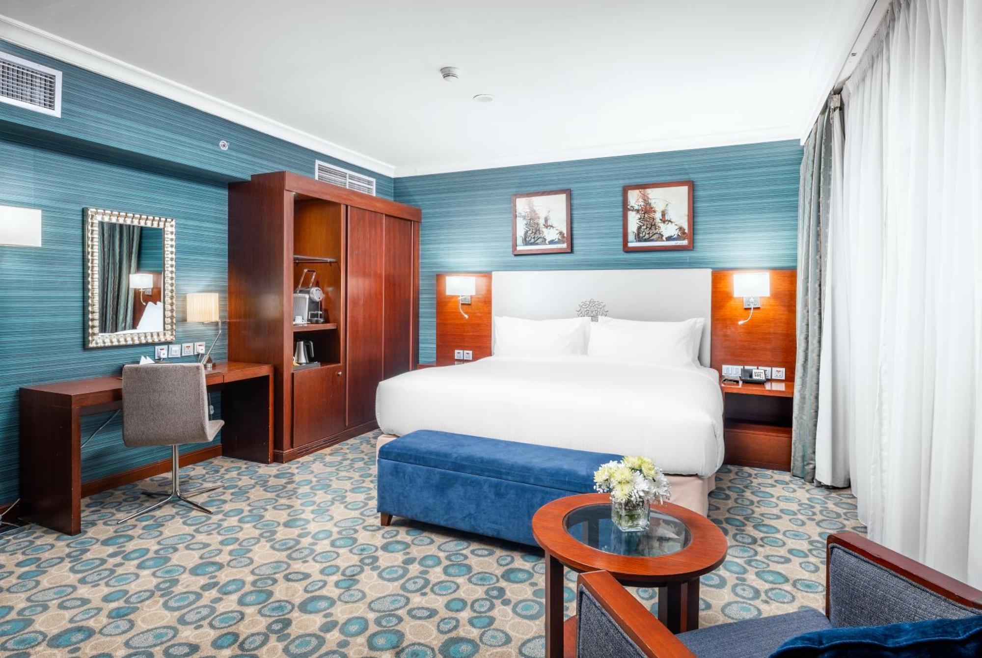 Al Aqeeq Madinah Hotel Médine Extérieur photo The photo shows a well-decorated hotel room. It features a large bed with white bedding, flanked by wall-mounted lights. Behind the bed, there are two framed pictures hanging on a blue wall. To the left, there's a wooden wardrobe and a desk with a ch