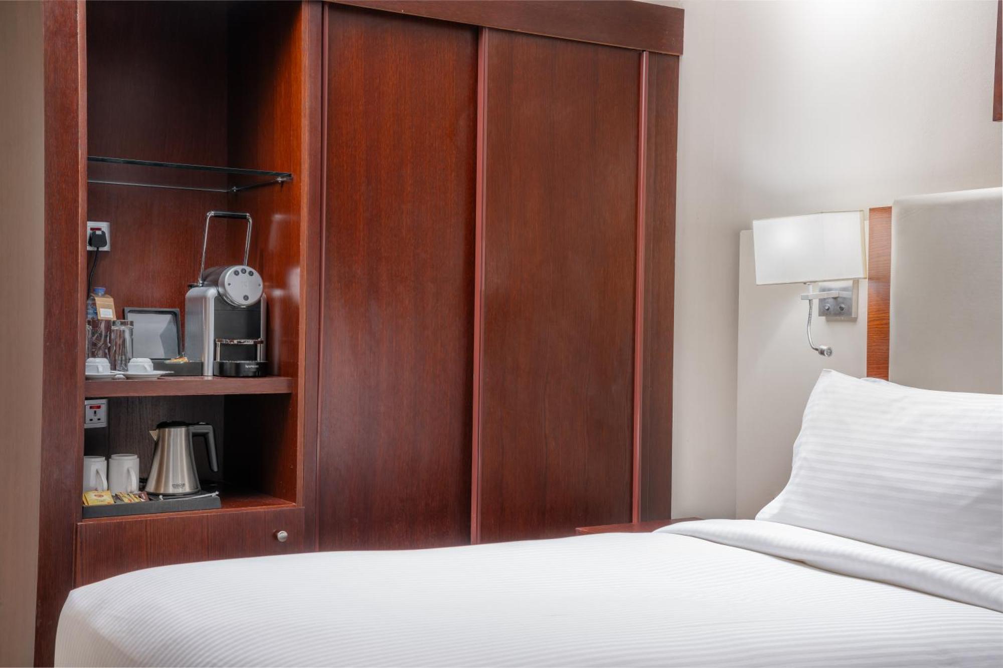 Al Aqeeq Madinah Hotel Médine Extérieur photo The photo shows a hotel room with a minimalist design. On the left side, there's a wooden cabinet with sliding doors that houses a small coffee maker and additional amenities, such as cups and a kettle. The bed is neatly made with white bedding and i