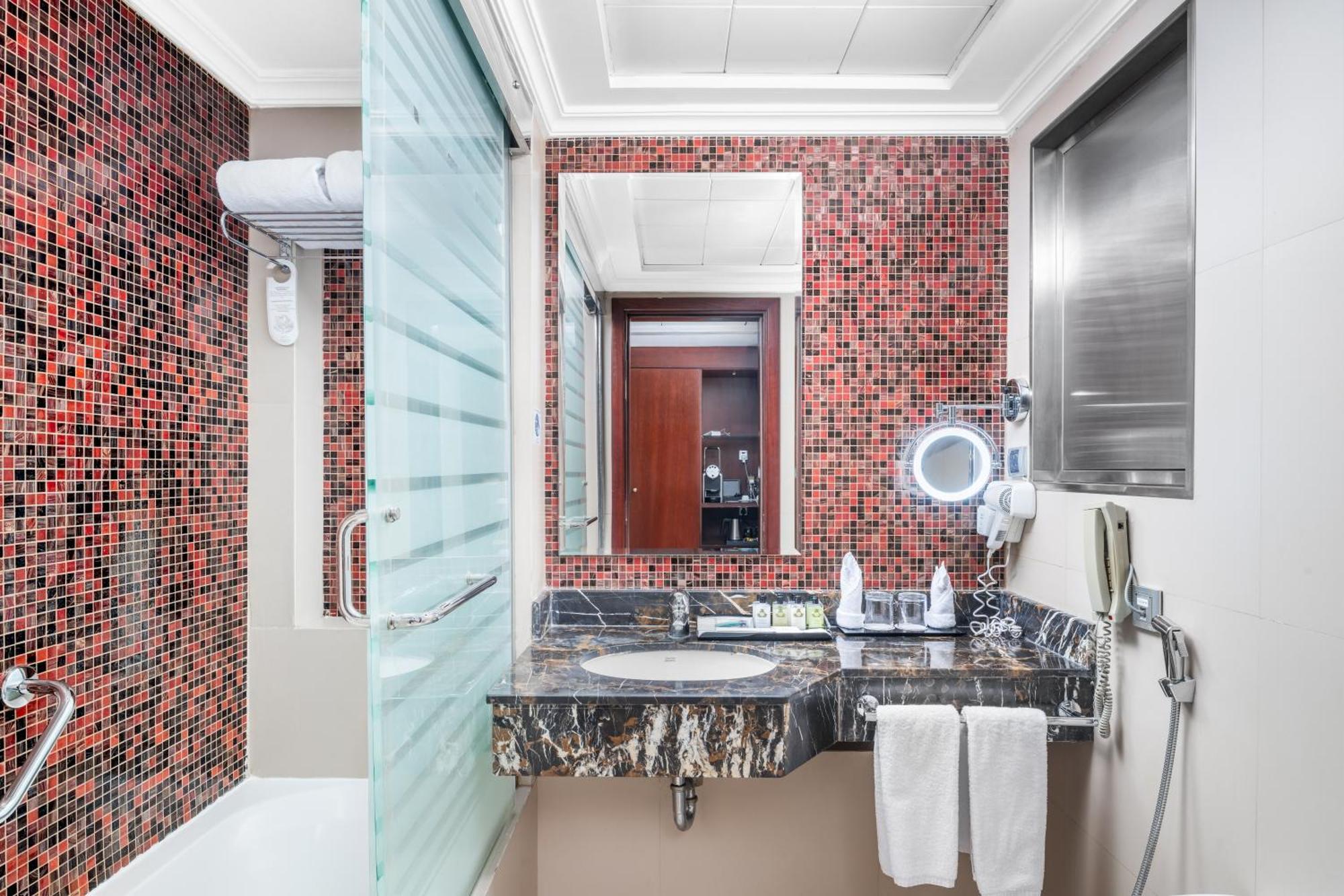 Al Aqeeq Madinah Hotel Médine Extérieur photo The photo shows a modern bathroom featuring a mix of design elements. The walls have a vibrant red mosaic tile pattern, adding a bold touch to the space. There is a glass shower enclosure, and next to it, a sleek countertop with a dark, marbled surfa