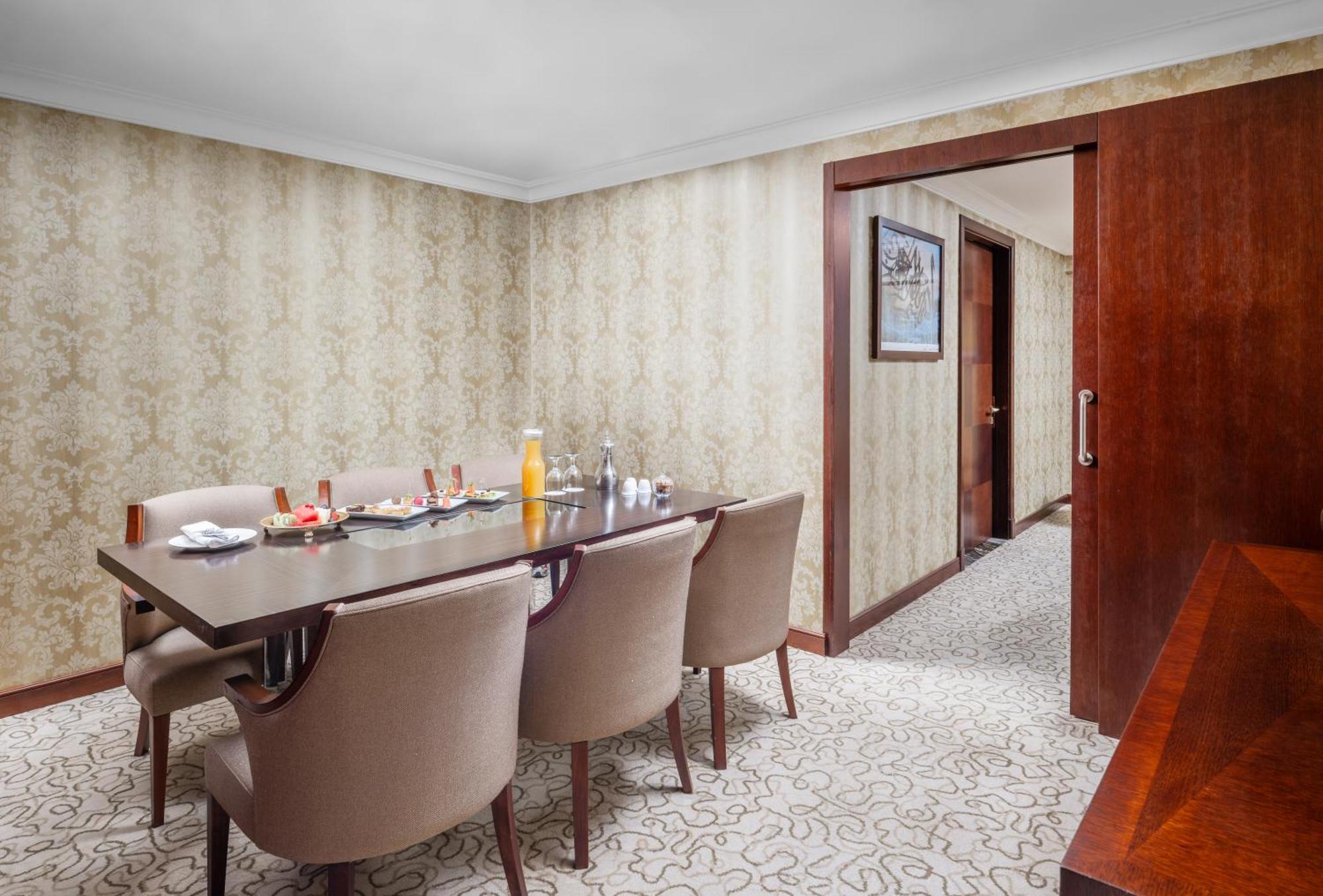 Al Aqeeq Madinah Hotel Médine Extérieur photo The photo shows a dining room with a stylish and elegant design. There is a large wooden dining table surrounded by plush, beige chairs. On the table, there are various items including a bottle of orange juice, glasses, and other dining accessories. 