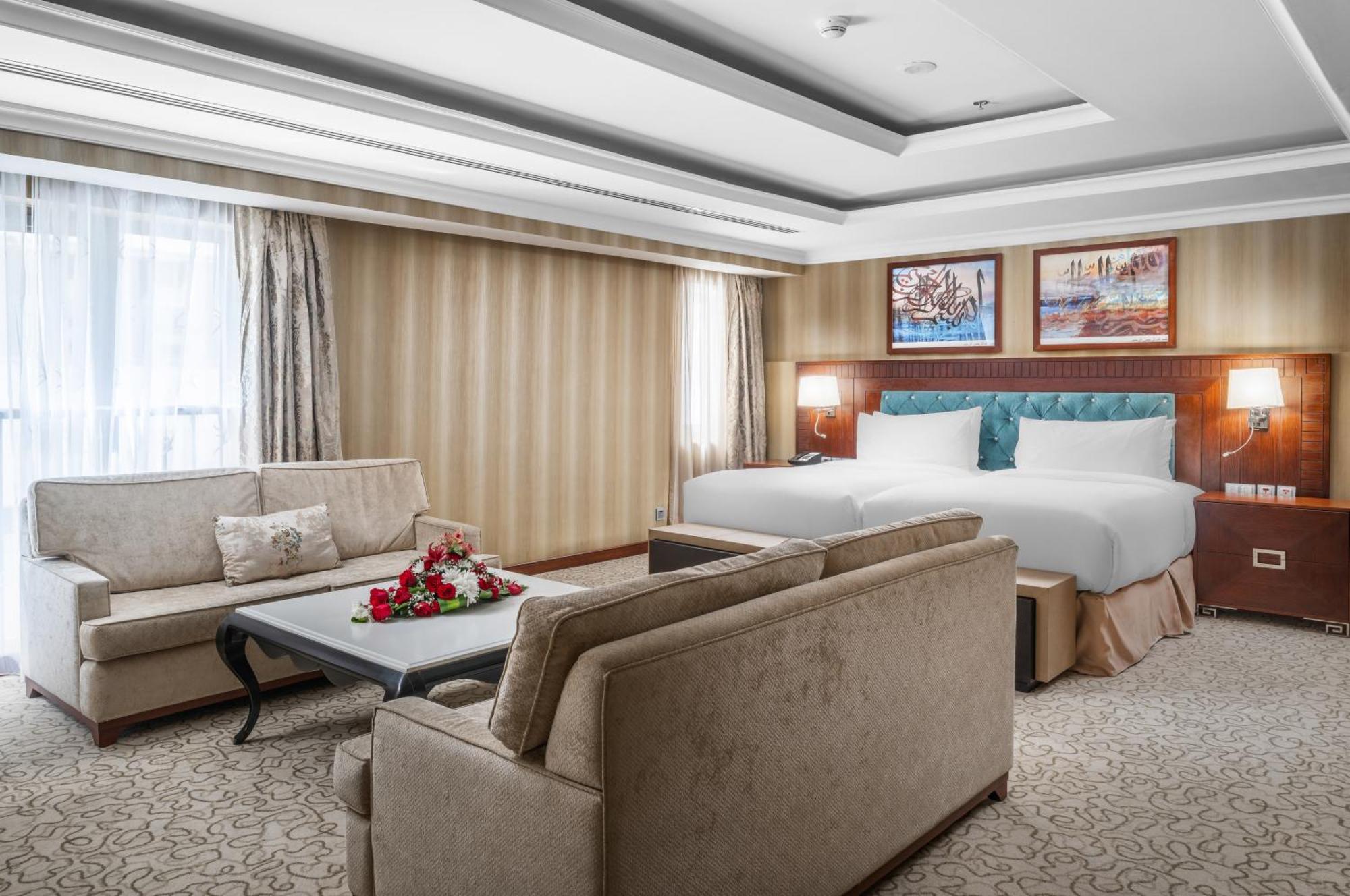Al Aqeeq Madinah Hotel Médine Extérieur photo The photo shows a well-decorated hotel room. It features a large bed with two pillows and a light-colored duvet. There are two comfortable couches arranged around a coffee table with a floral centerpiece. The walls have a subtle striped design, and l