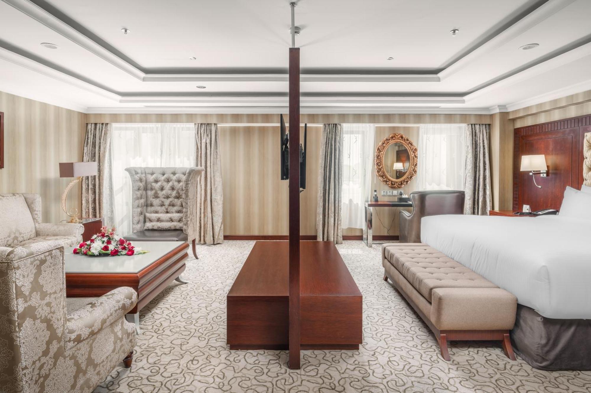 Al Aqeeq Madinah Hotel Médine Extérieur photo The photo showcases a spacious hotel suite with an elegant and modern design. The room features a large bed on one side, complemented by a plush bench at the foot. Opposite the bed, there is a stylish seating area that includes a decorative armchair 