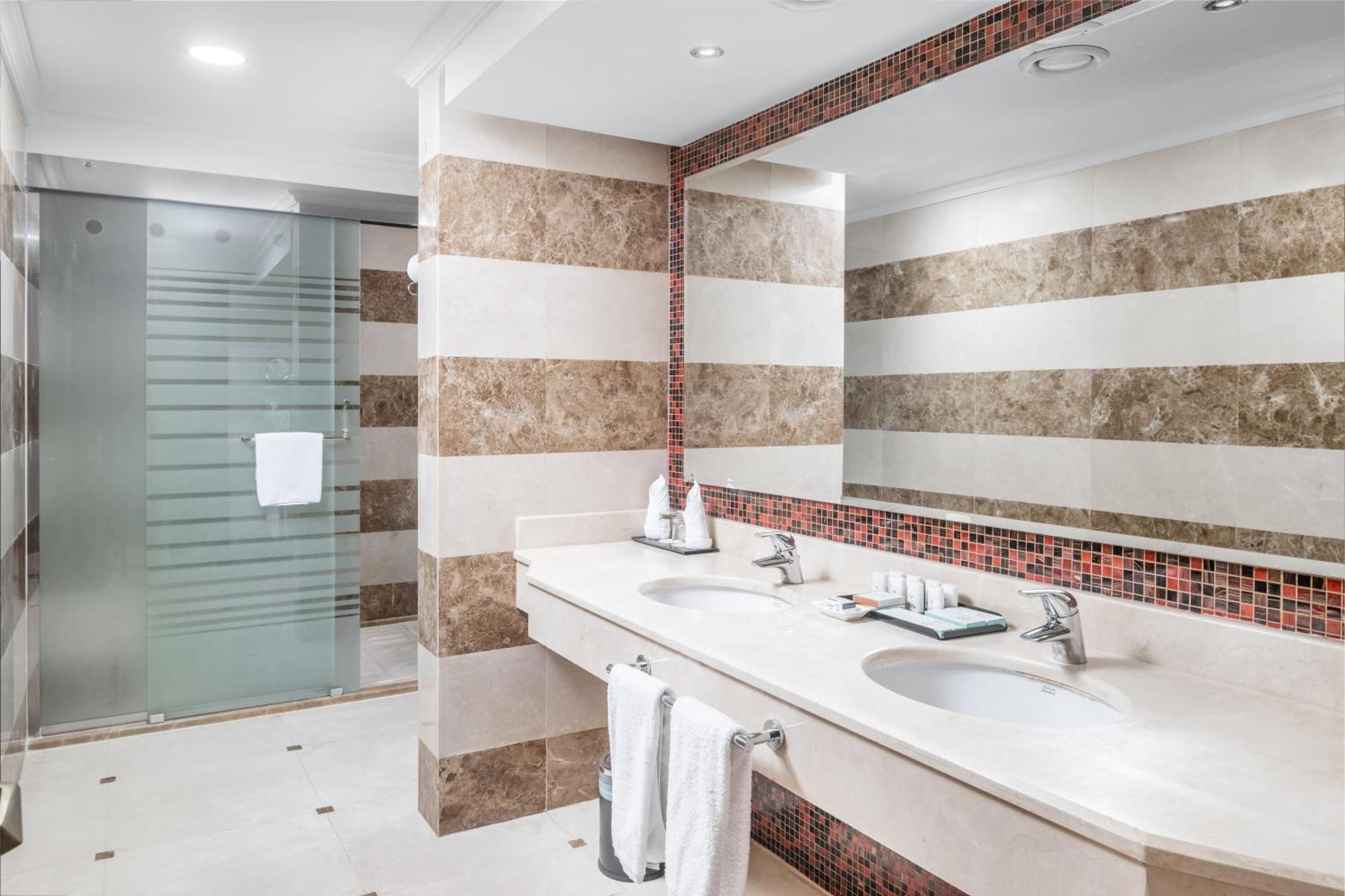 Al Aqeeq Madinah Hotel Médine Extérieur photo The photo shows a modern bathroom with an elegant design. It features a large double sink with a smooth countertop, accompanied by faucets. The walls are adorned with alternating horizontal stripes of light and dark materials, creating a stylish look