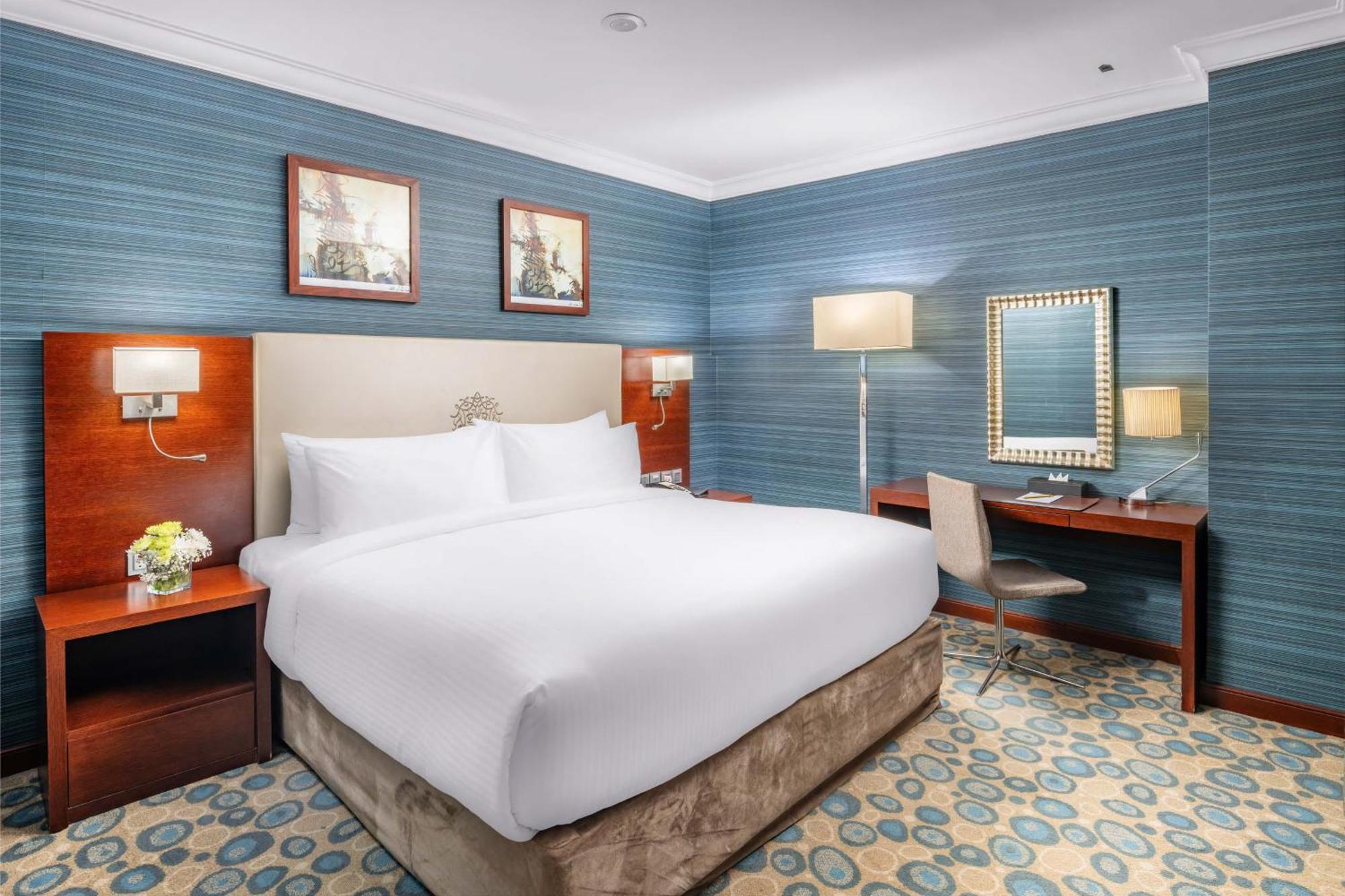 Al Aqeeq Madinah Hotel Médine Extérieur photo The photo shows a well-furnished hotel room. It features a large bed with white linens, flanked by wooden nightstands. The walls are painted in a light blue color with a textured pattern. Above the bed, there are two framed pictures. On one side of t
