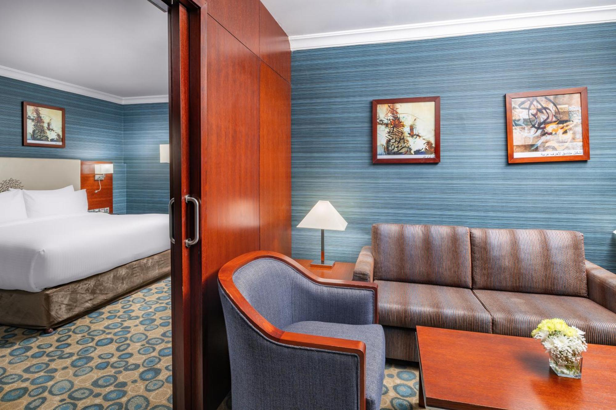 Al Aqeeq Madinah Hotel Médine Extérieur photo The photo shows a well-furnished hotel room featuring a living area and a bedroom. To the left, there is a bed visible through an open doorway. The living area includes a gray upholstered chair and a brown couch, both arranged around a small table. A