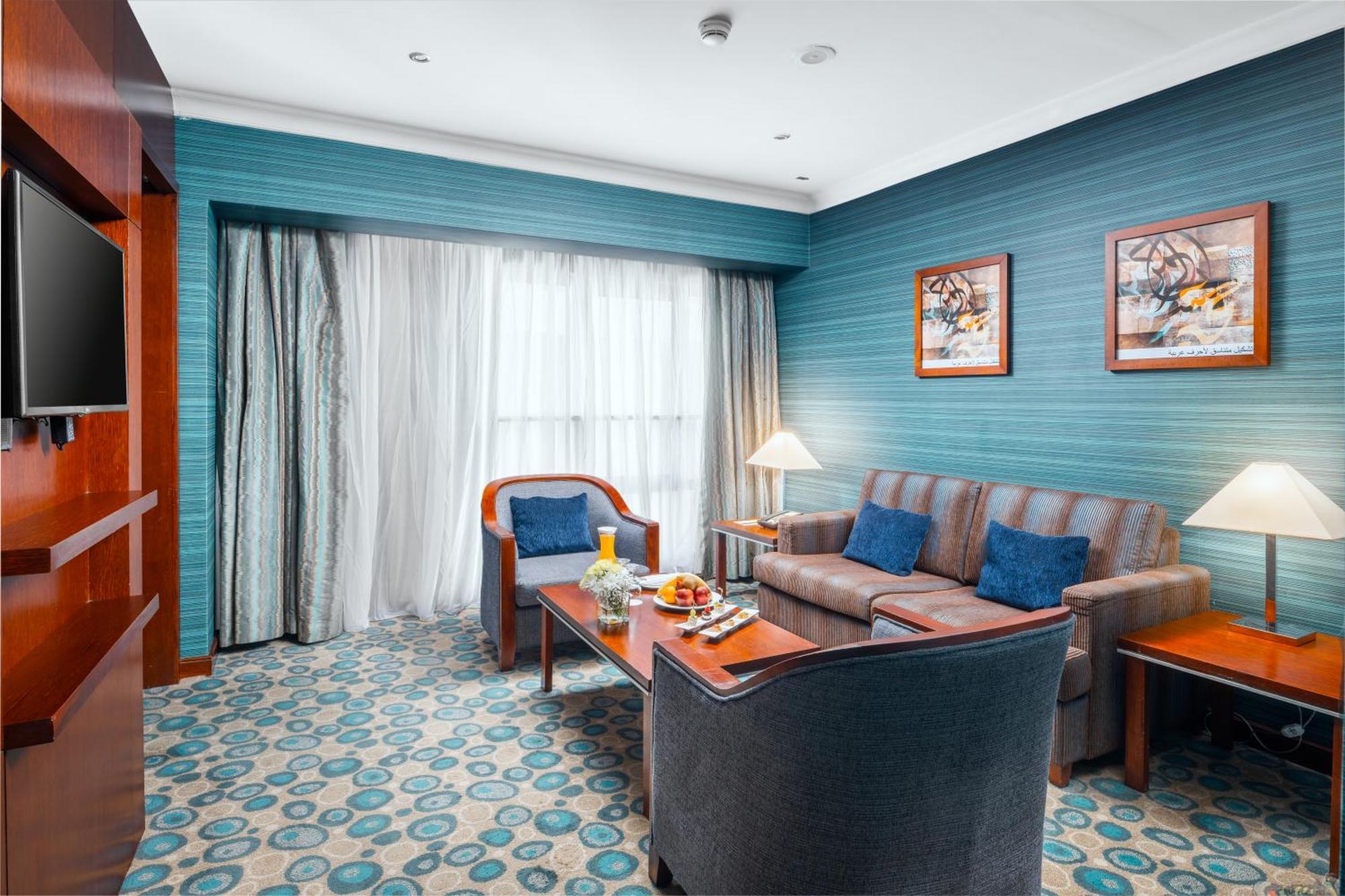Al Aqeeq Madinah Hotel Médine Extérieur photo The photo shows a cozy and modern hotel room featuring a sitting area. The walls are painted a calming teal color, complemented by a patterned carpet in shades of blue and beige. There is a comfortable sofa with cushions, two armchairs, and a wooden 
