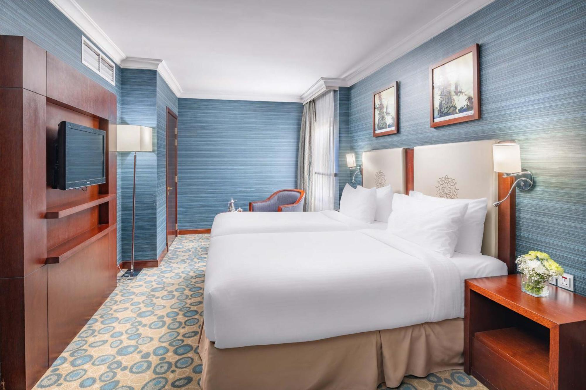 Al Aqeeq Madinah Hotel Médine Extérieur photo The photo shows a hotel room featuring two twin beds dressed in white linens. The walls are painted in a soft blue color, and there are framed artworks hung above the beds. A flat-screen TV is mounted on a wooden unit against one wall. To the side, t