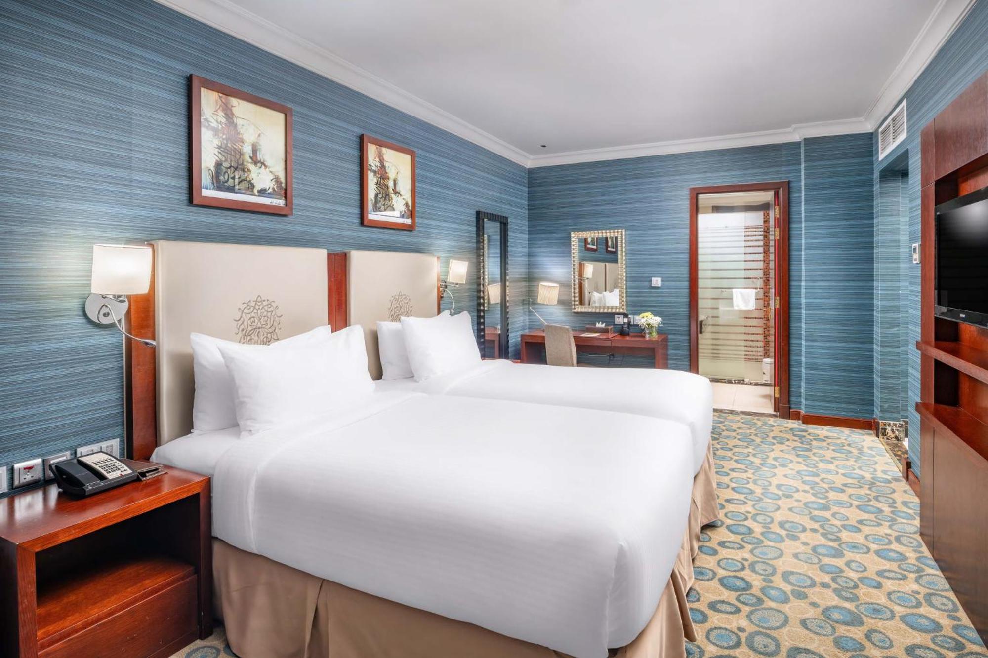 Al Aqeeq Madinah Hotel Médine Extérieur photo The photo shows a hotel room featuring two twin beds with white bedding, positioned against a light-colored headboard. The walls are painted in a soft blue shade, adorned with framed artwork. There is a wooden desk with a chair, and a mirror mounted 