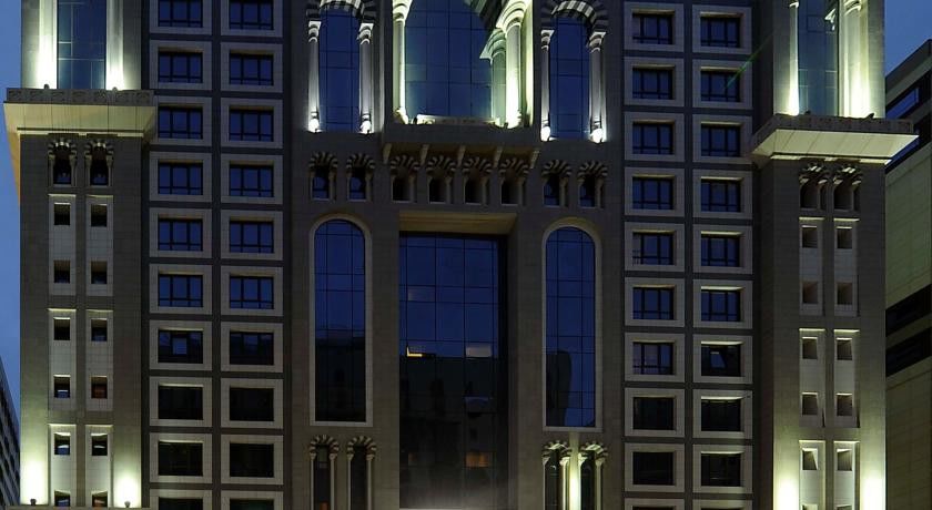 Al Aqeeq Madinah Hotel Médine Extérieur photo The image shows the facade of a modern high-rise building illuminated at night. The design features large glass windows arranged in a grid pattern, along with decorative architectural elements at the top, including arches and ornate detailing. The co