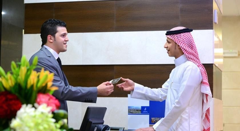 Al Aqeeq Madinah Hotel Médine Extérieur photo The image depicts two individuals at a reception desk. The person on the left, dressed in a formal suit, appears to be handing a card or document to the person on the right, who is wearing traditional attire, including a white thobe and a red and whi