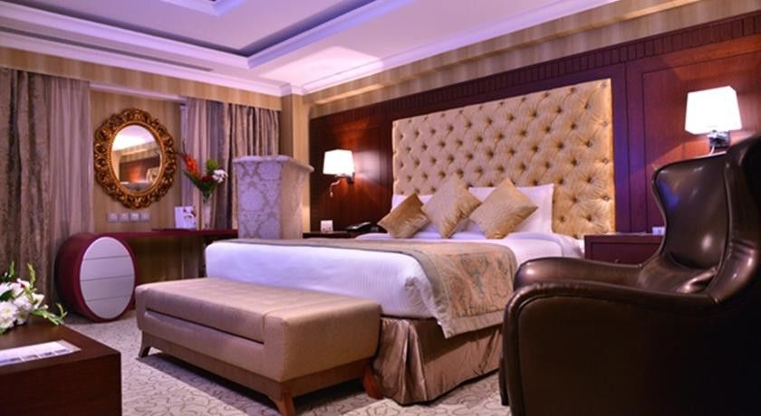 Al Aqeeq Madinah Hotel Médine Extérieur photo The photo shows a luxurious hotel room. It features a large, upholstered headboard with a tufted design, paired with plush bedding and decorative cushions. There's a stylish bench at the foot of the bed and a comfortable armchair to the side. The roo