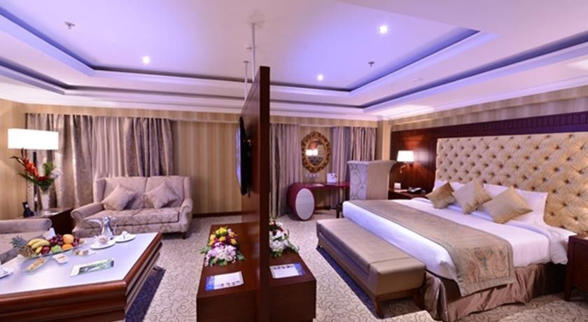 Al Aqeeq Madinah Hotel Médine Extérieur photo The image shows a luxurious hotel room. It features a large bed with a tufted headboard, flanked by nightstands with lamps. There's a seating area with a plush sofa and a coffee table. A decorative partition separates the bed from the living area. Th