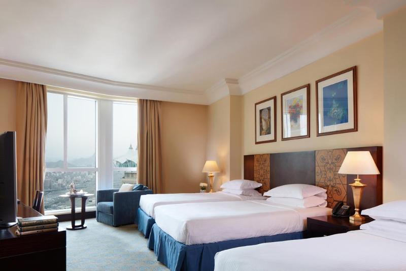 Al Aqeeq Madinah Hotel Médine Extérieur photo The photo shows a hotel room featuring three beds arranged neatly. The room has a warm, inviting ambiance with beige walls and a soft carpet. Large windows allow natural light to fill the space, offering a view outside. There are curtains in a light 