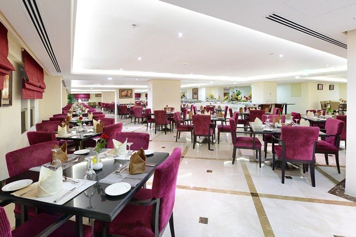 Al Aqeeq Madinah Hotel Médine Extérieur photo The photo shows an elegant restaurant interior with a spacious layout. The dining area features multiple tables, each set with white tablecloths and dinnerware. The chairs are upholstered in a rich purple fabric, adding a touch of luxury to the atmos