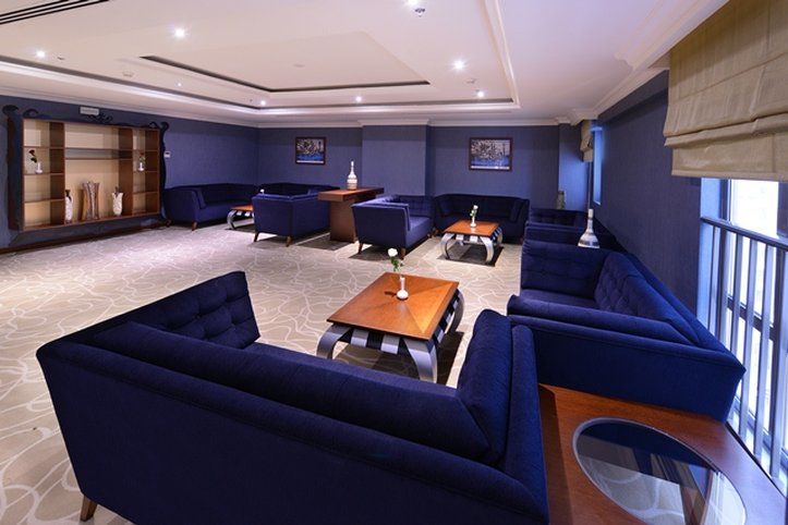 Al Aqeeq Madinah Hotel Médine Extérieur photo The photo shows a modern lounge area featuring a sophisticated design. The room is painted in a deep blue color, creating a calm and elegant atmosphere. There are several dark blue sofas arranged around a central wooden coffee table. The table is dec
