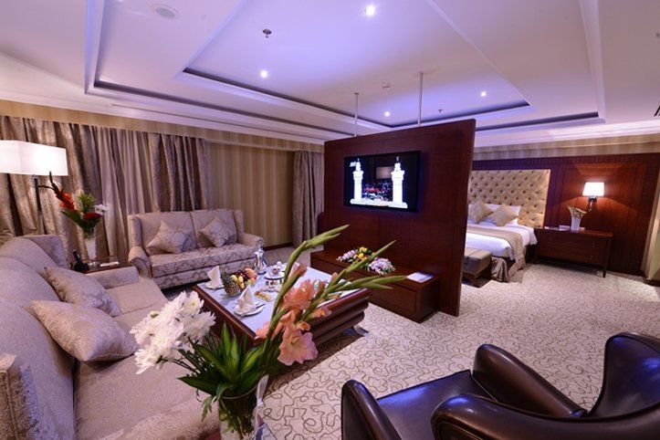 Al Aqeeq Madinah Hotel Médine Extérieur photo The photo shows a luxurious hotel room suite. There is a comfortable seating area with stylish sofas and a coffee table. A decorative arrangement of flowers is on the table, adding a touch of elegance. A large flat-screen television is mounted on a p
