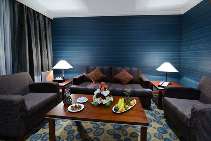 Al Aqeeq Madinah Hotel Médine Extérieur photo The photo shows a cozy living area featuring a dark blue wall with a textured design. In the center, there is a wooden coffee table surrounded by two comfortable armchairs and a sofa with decorative cushions. On the table, there are plates with snack