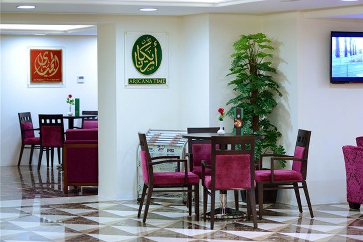 Al Aqeeq Madinah Hotel Médine Extérieur photo The photo shows an interior space, likely a dining or lounge area. There are several tables with four chairs each, featuring a modern design with dark wood and plush maroon upholstery. On the walls, there are decorative signs, one of which has Arabic