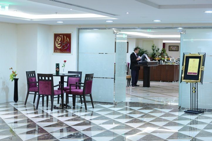 Al Aqeeq Madinah Hotel Médine Extérieur photo The photo shows a modern interior space, likely a lobby or reception area of a hotel or restaurant. It features a checkered floor with a combination of dark and light tiles. On the left, there is a small table set for two, adorned with a single rose 