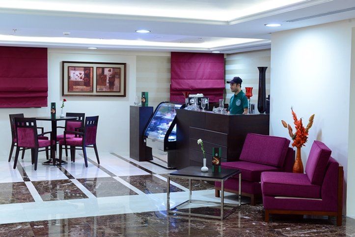 Al Aqeeq Madinah Hotel Médine Extérieur photo The photo shows a modern interior space, likely a café or small restaurant. It features a seating area with a few tables and chairs, all in a dark wood finish with purple upholstery. There is a counter area with a staff member behind it, possibly ser