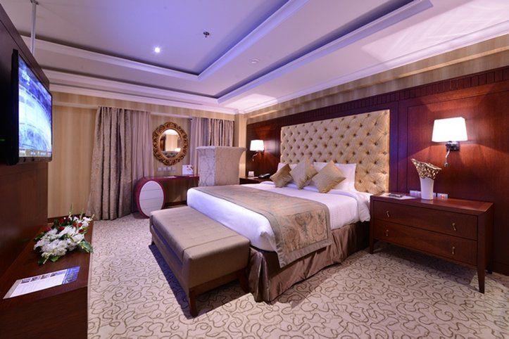 Al Aqeeq Madinah Hotel Médine Extérieur photo The photo shows an elegantly designed hotel room. It features a large bed with a plush headboard and decorative pillows. On either side of the bed are bedside tables with lamps. There's a comfortable bench at the foot of the bed and a TV mounted on t