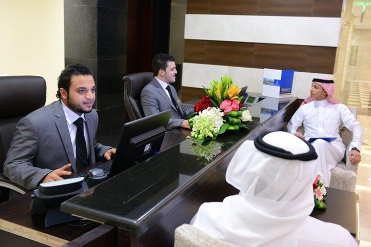 Al Aqeeq Madinah Hotel Médine Extérieur photo The photo depicts a reception area with three individuals. Two men are seated behind a reception desk; they are dressed in formal suits and appear to be engaged in conversation, possibly assisting a guest. The desk has a computer and a vibrant arrang