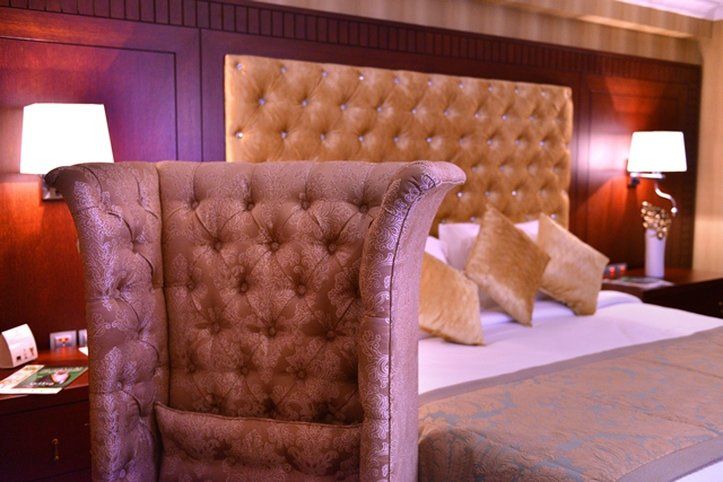 Al Aqeeq Madinah Hotel Médine Extérieur photo The image shows a luxurious bedroom featuring a large upholstered headboard with a tufted design in a soft, beige color. The bed is elegantly dressed with decorative pillows and a textured bedspread. In the background, there are wooden paneling and s